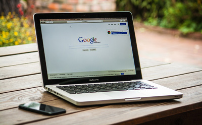Laptop steht auf Holztisch im Freien und zeigt mit offenem Browser Google-Seite an; danebenliegend ein Smartphone